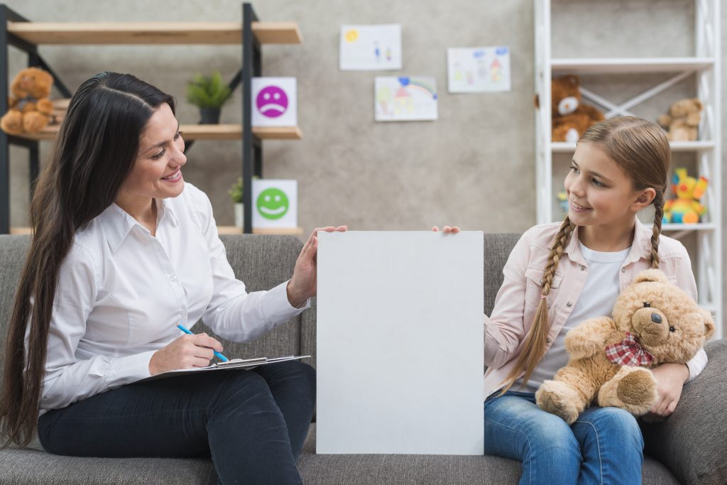 Friendly psychologist and little girl holding blank placard sitting on sofa
