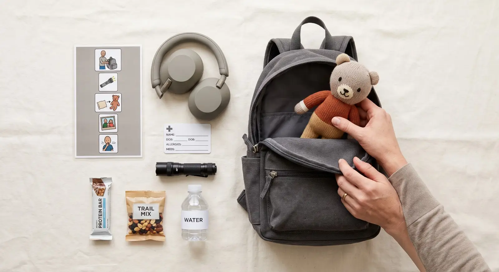 A parent's hands arrange the contents of a small emergency preparedness bag — noise-canceling headphones, a laminated picture card, a comfort toy, a medical information card, a flashlight, and snacks — in a calm, organized flat-lay.