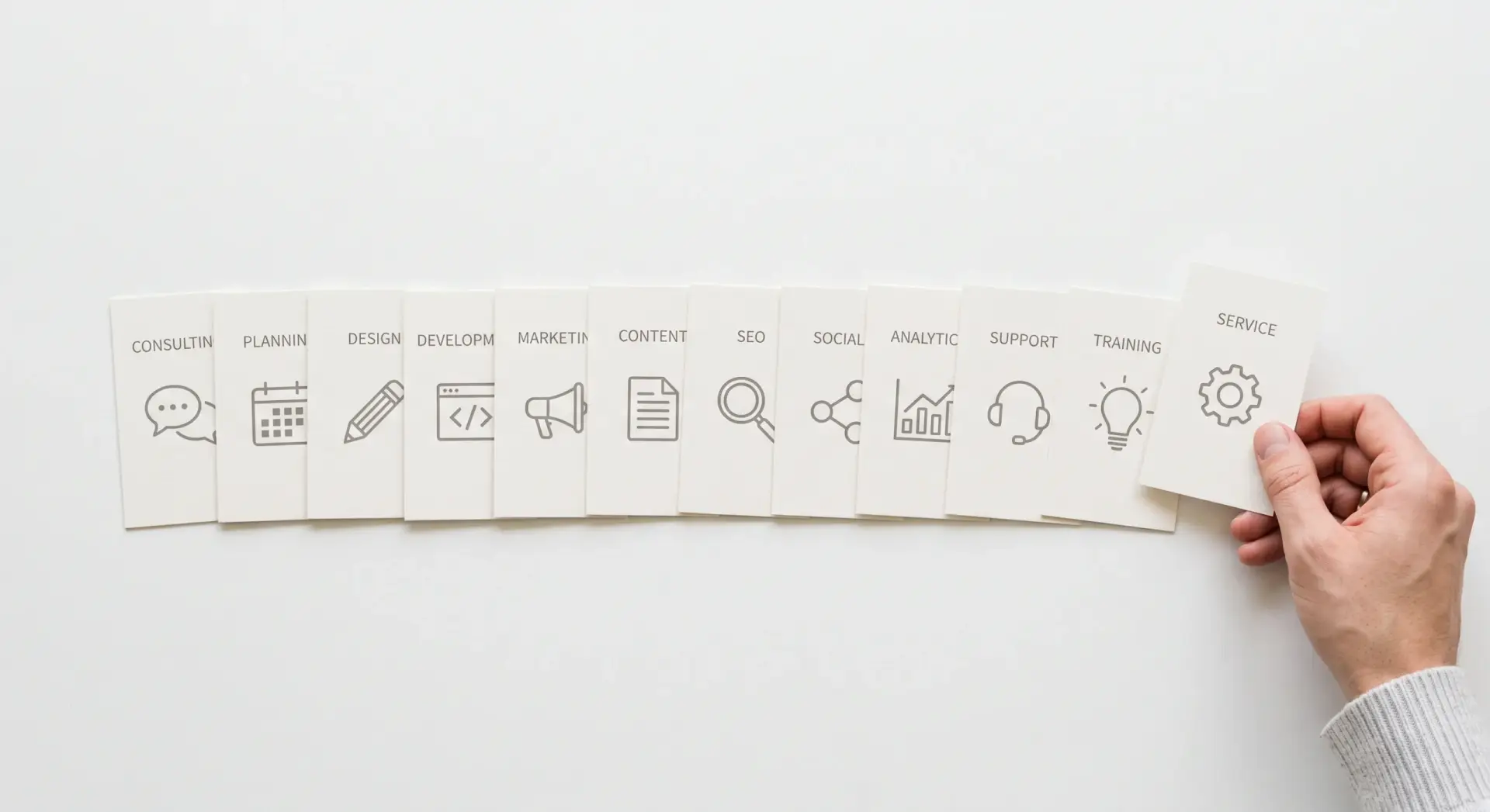 A clean, near-straight-down flat-lay photograph of a row of simple printed service icon-cards fanned out on a plain white surface, a hand laying the last card into place. Even, bright, high-key lighting, minimal shadow. Neutral palette — white and pale grey with soft muted line-art icons, no saturated color. Clear, tidy, informational mood. No readable brand text or logos.