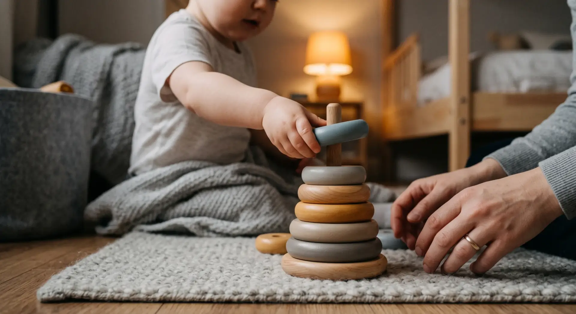 A candid photograph of a young white child stacking wooden rings on a bedroom floor, a parent's out-of-focus hands nearby ready to help. Close crop from a slightly elevated angle on the child's hands and the toy, no faces in frame. Shallow depth of field. Warm, cozy, low-key ambient light from a small table lamp in the late afternoon. Muted palette of soft grey with a warm honey-wood tone as the accent. Playful, gentle, unposed. No text, no logos, no harsh shadows; keep top corners uncluttered.