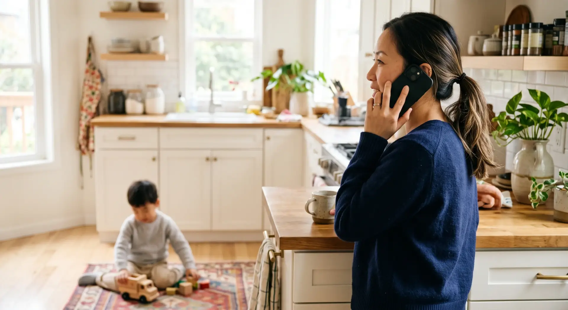 A candid photograph of an East Asian parent standing at a kitchen counter holding a phone to their ear, a young child playing with a toy on the floor softly blurred in the foreground. Over-the-shoulder vantage past the parent, whose face is in partial profile. Shallow depth of field. Bright, clear kitchen daylight, crisp and even. Muted palette of warm white and pale wood with a navy accent (the parent's top). Everyday, candid, quietly hopeful. No text, no logos, no harsh shadows; leave the top corners clear.
