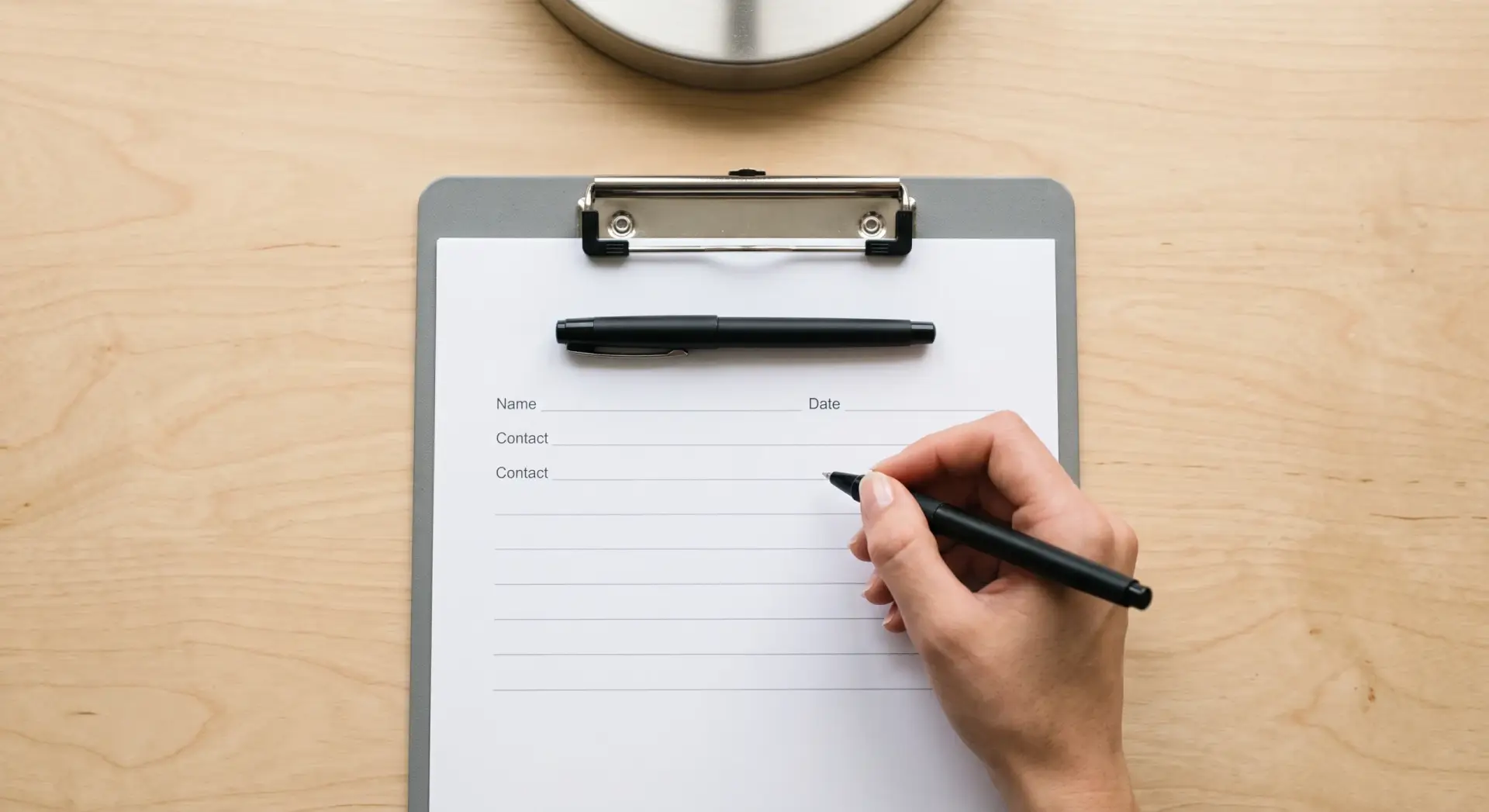 A clean, near-straight-down flat-lay photograph of a blank intake form on a clipboard with a pen laid across it, a hand poised to begin writing. Bright, direct light from an overhead desk lamp — clean and functional, minimal soft shadow. Neutral palette of white paper, grey clipboard and black pen. Simple, approachable, get-started mood. No readable brand text or logos.