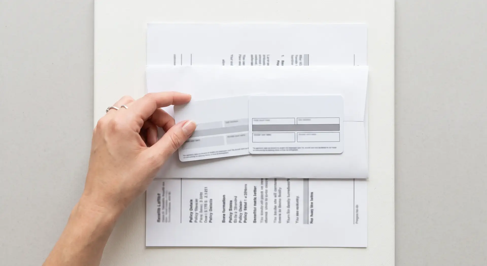A clean, straight-down flat-lay photograph of two generic insurance cards set on top of a folded benefits letter, a hand sliding one card into view. Even, bright, high-key lighting, minimal shadow. Neutral palette of white and cool grey with plain unbranded cards; any text indistinct. Clear, factual, reassuring-informational mood. No readable brand text or logos.